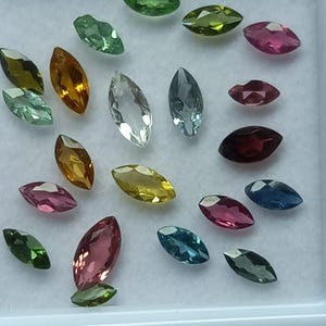 Pode incluir: Uma coleção de pedras preciosas com corte marquise em várias cores, incluindo verde, amarelo, rosa, azul e transparente. As pedras preciosas são exibidas numa bandeja branca, mostrando as suas superfícies polidas e tons vibrantes. As pedras são de vários tamanhos.