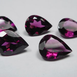 Pode incluir: Cinco pedras preciosas facetadas em forma de pêra, em tons de roxo profundo e magenta. As gemas são cortadas para refletir a luz, mostrando sua estrutura interna e variações de cor. As pedras estão dispostas sobre uma superfície branca.