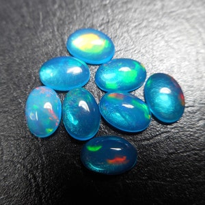 以下が含まれることがあります： 青、緑、赤の色合いが美しい8つの楕円形のオパール。宝石は虹色の輝きを持ち、色の遊びを作り出しています。オパールは暗く、質感のある背景に配置されています。