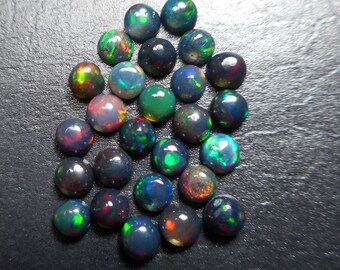 May include: A collection of round, polished black opals with vibrant flashes of color. The opals display a range of hues, including green, red, and blue, creating a dazzling effect. The gemstones are scattered on a dark surface.
