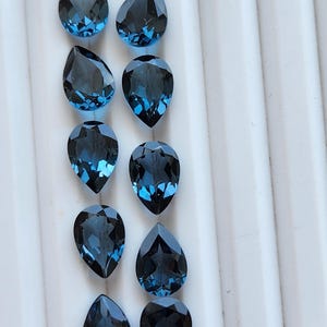 Pode incluir: Dez pedras preciosas facetadas em forma de pera de uma cor azul profunda e saturada. As pedras estão dispostas em duas colunas verticais de cinco, cada uma exibindo facetas geométricas complexas que refletem a luz nas suas superfícies polidas de azul escuro.