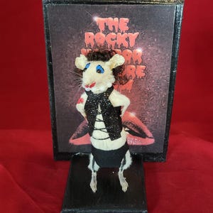 May include: A taxidermied rat dressed as Dr. Frank-N-Furter from the movie "The Rocky Horror Picture Show". The rat is wearing a black leather jacket, a white shirt, and black pants. The rat is standing on a black base in front of a framed poster of the movie.