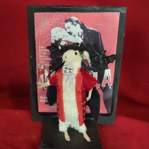 May include: A whimsical Halloween-themed figurine of a white mouse dressed as a vampire, complete with a red jacket, skull-shaped bow tie, and bat wings. The mouse holds a miniature glass of red liquid, set against a vintage movie poster backdrop.