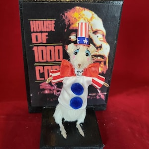 May include: A taxidermied white mouse wearing a red, white, and blue clown costume with a skull and crossbones on its chest. The mouse is standing on a black base in front of a poster for the movie "House of 1000 Corpses."
