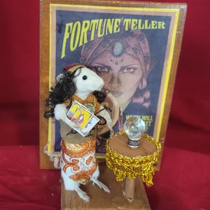 May include: A whimsical figurine of a white rat dressed as a fortune teller, holding tarot cards. The rat stands before a vintage "Fortune Teller" poster and a small table with a crystal ball. The rat wears a turban, shawl, and patterned skirt.