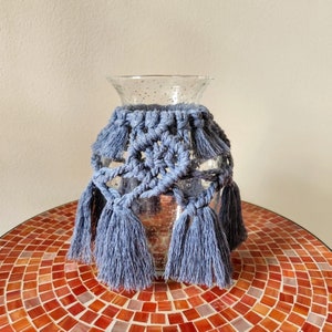 May include: A clear glass vase with a textured surface is decorated with a blue crocheted macrame cover with tassels.