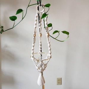 May include: A white macrame plant hanger with wooden beads and a gold metal ring. The hanger is hanging from a hook on a white wall.