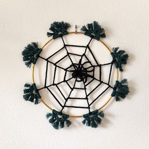 May include: A black macrame spider web design with a gold metal hoop and dark green tassels hanging from the hoop.
