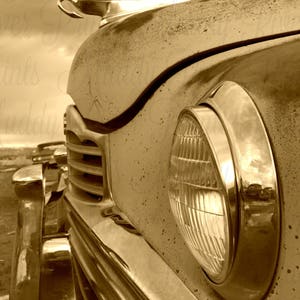 May include: Close-up of a vintage car's chrome headlight and hood ornament. The car is a light brown color and has a weathered, aged look. The headlight is round and has a chrome trim. The hood ornament is a winged figure.