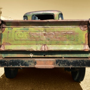 May include: Rear view of a vintage Chevrolet pickup truck. The truck bed is weathered green with the embossed word "CHEVROLET". The bumper and fenders show signs of rust and age, with a black bumper and a single taillight visible.