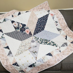 May include: A handmade quilt with a geometric starburst design. The quilt features various fabrics in white, pink, navy, gray, and floral patterns. The border is a light pink with a gold leaf pattern. The quilt is folded on a gray sofa.