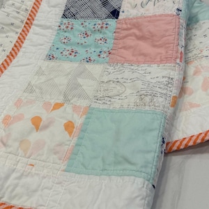 May include: A patchwork quilt with a white background and various colored squares. The squares are made of different fabrics with patterns including floral, geometric, and text. The quilt has an orange and white striped border.