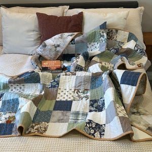 May include: A patchwork quilt with squares of blue, green, and white fabrics, some with floral patterns, lies on a bed with white pillows and a burgundy pillow. The book "Where the Crawdads Sing" is visible. The quilt has a brown border.