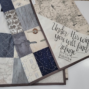May include: A patchwork quilt with a brown border. The quilt is made of various fabrics with different patterns, including maps, airplanes, and geometric designs. The quilt has a white panel with the text "Under His wings you will find refuge PSALM 91:4" written in black.