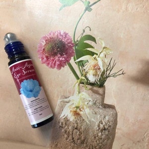 May include: A blue glass roller bottle with a white label that reads "Anti-Aging Eye Serum". The bottle is sitting on a light brown surface with a vase of flowers in the background.