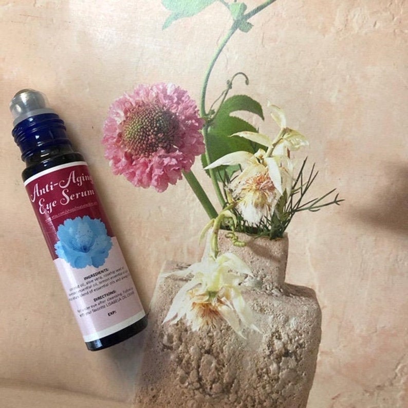 May include: A blue glass roller bottle with a white label that reads "Anti-Aging Eye Serum". The bottle is sitting on a light brown surface with a vase of flowers in the background.