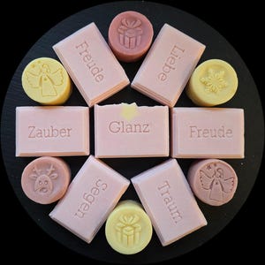 Puede incluir: Una colección de jabones hechos a mano en varias formas y colores. Jabones rectangulares con palabras alemanas en relieve como "Freude" y "Liebe" se organizan alrededor de jabones circulares con diseños de ángeles, regalos y copos de nieve. Los jabones son rosas y amarillos.