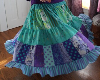 May include: A woman wearing a purple top and a multi-colored patchwork skirt with ruffles. The skirt is made of different fabrics and patterns, including polka dots, stripes, and floral prints.