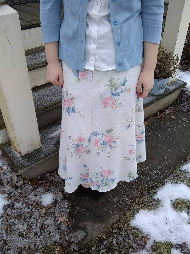 May include: A white skirt with a floral pattern, worn with a light blue cardigan and a white button-down shirt.