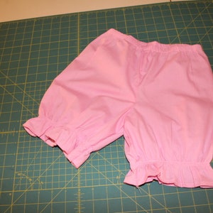 May include: Pink cotton bloomers with a ruffled hem. The bloomers are made of a soft, lightweight fabric and have a comfortable elastic waistband.