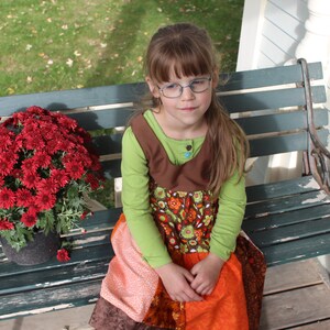 May include: A young person wearing glasses and a colorful dress sits on a green bench. A large pot of red chrysanthemums sits to the left. The dress has a green top, brown bodice, and an orange and patterned skirt.