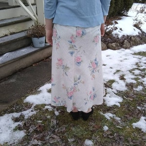 May include: A long white skirt with a floral print featuring pink, blue, and green flowers. The skirt is worn with a light blue sweater.