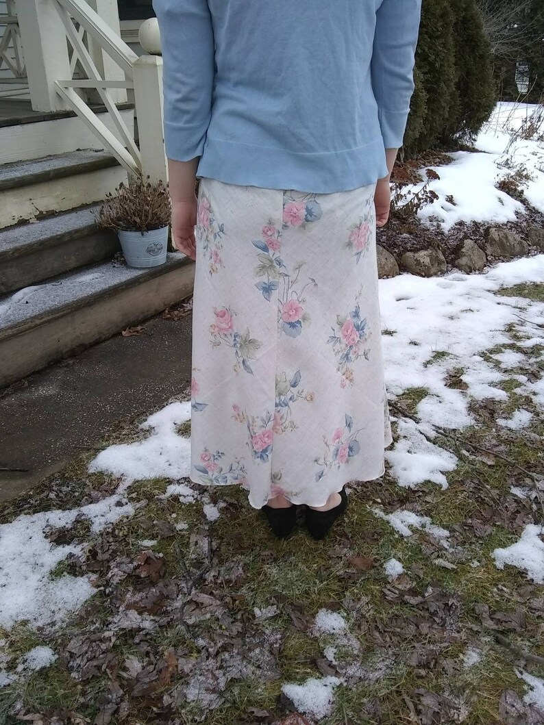 May include: A long white skirt with a floral print featuring pink, blue, and green flowers. The skirt is worn with a light blue sweater.