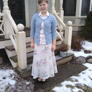 May include: A woman wearing a light blue cardigan over a white button-down shirt and a floral print skirt. She is standing on a porch with snow on the ground.