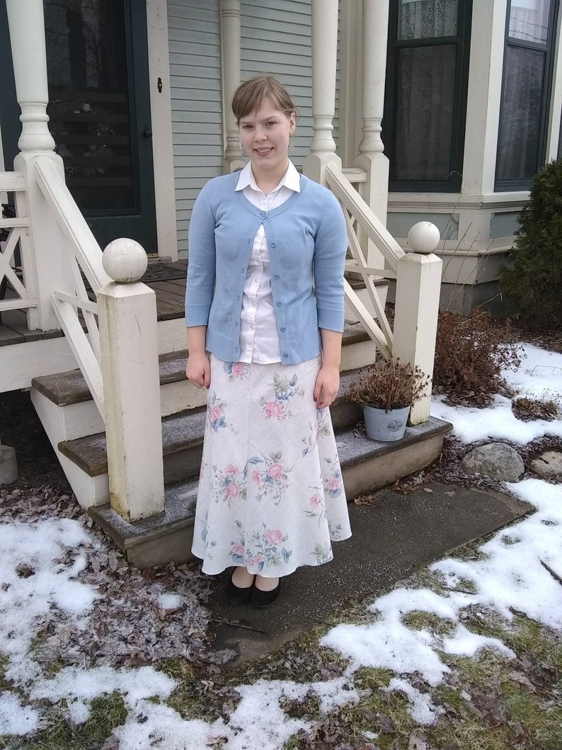 May include: A woman wearing a light blue cardigan over a white button-down shirt and a floral print skirt. She is standing on a porch with snow on the ground.