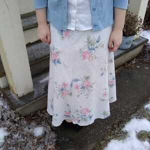 May include: A white floral skirt with a blue cardigan and white button-down shirt. The skirt has a light blue and pink floral pattern.