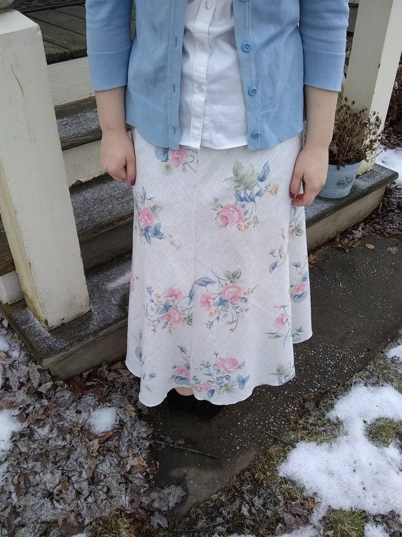 May include: A white floral skirt with a blue cardigan and white button-down shirt. The skirt has a light blue and pink floral pattern.