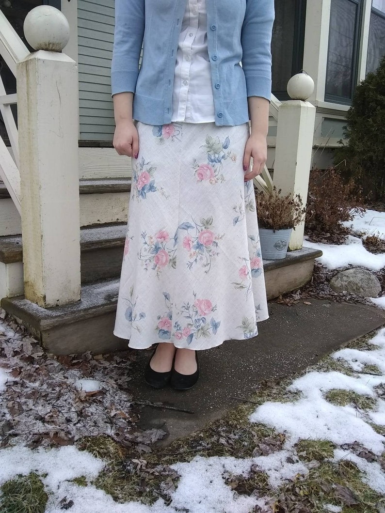 May include: A woman wearing a light blue cardigan over a white button-down shirt and a white floral skirt. She is standing on a set of stairs with snow on the ground.