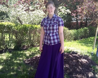 May include: A young person wearing a purple skirt and a plaid shirt with a white background and black and purple squares. The person is standing in front of a tree with purple flowers.