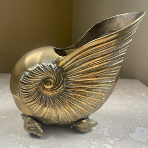 May include: A brass-colored decorative nautilus shell-shaped vase. The vase has a detailed spiral design on the body and a flared opening. It rests on three small, decorative feet.