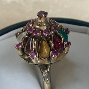 May include: A gold-toned, ornate jewelry piece with a crown-like design. The crown is adorned with various gemstones, including red, green, and yellow stones, and smaller pink gems. The piece has a long, slender handle.