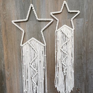 May include: Two white macrame star wall hangings with a geometric pattern. The stars are made of white cotton cord and have a fringe at the bottom.