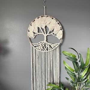 May include: A white macrame wall hanging in the shape of a tree with a wooden hoop. The tree has leaves and roots and is hanging from a hook on a grey wall. A green plant is in the foreground.