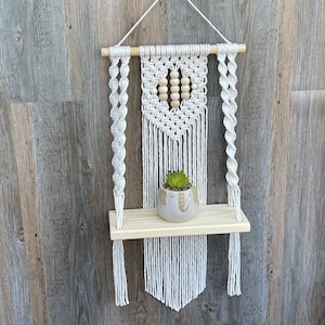 May include: White macrame wall hanging with a wooden shelf and a small potted succulent. The macrame features a heart shape with wooden beads.