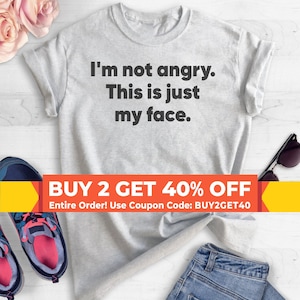 May include: A gray t-shirt with the text "I'm not angry. This is just my face." printed on it. The shirt is laying on a white surface with a pair of blue jeans, a pair of sneakers, and sunglasses. The text "BUY 2 GET 40% OFF" is printed on a red and yellow banner.