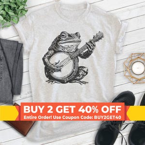 May include: A gray t-shirt with a black and white illustration of a frog playing a banjo. The text "BUY 2 GET 40% OFF" is printed in red and yellow on a white banner. The text "Entire Order! Use Coupon Code: BUY2GET40" is printed in black below the banner.