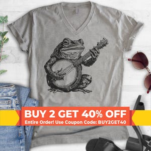 May include: A heather gray V-neck t-shirt featuring a black ink-style illustration of a frog playing a banjo. The graphic is centered on the chest. The shirt has short sleeves and a standard fit. The text BUY 2 GET 40% OFF is printed across.
