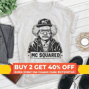 May include: A light gray short-sleeve t-shirt featuring a black graphic print of a person with curly hair, glasses, and a chain necklace, wearing a cap labeled MC2. Below the figure, the text MC SQUARED is printed in a bold, black rectangular box.