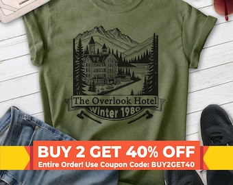 May include: An olive green t-shirt featuring a black graphic print of a large hotel nestled in a mountain landscape with pine trees. The text reads The Overlook Hotel winter 1980, accompanied by two crossed axes at the bottom of the design.