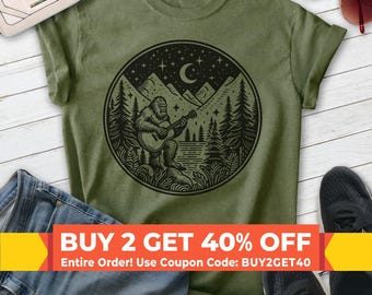 Op de afbeelding: Een olijfgroen t-shirt met een cirkelvormige zwarte afbeelding van een wezen dat op Bigfoot lijkt, spelend op een akoestische gitaar in een bosrijke omgeving met bergen, een maansikkel en sterren op de achtergrond, gecentreerd op de borst.