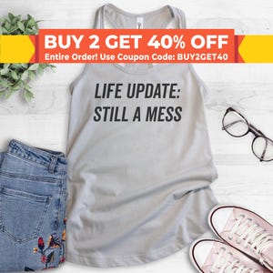 May include: A light gray tank top with the text "LIFE UPDATE: STILL A MESS" printed in black. The tank top is photographed on a white wooden surface with a pair of blue jeans, a pair of glasses, and two pairs of white sneakers.