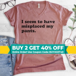 May include: A dusty rose V-neck t-shirt with the phrase "I seem to have misplaced my pants" in black lettering. The shirt is displayed with a pair of jeans, glasses, and sneakers. A promotional banner is at the bottom.