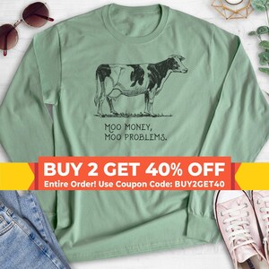 May include: A sage green long-sleeve shirt featuring a black line-art illustration of a cow standing in grass. Below the cow, the text reads: MOO MONEY, MOO PROBLEMS. The shirt is displayed with sunglasses, blue denim fabric, and white sneakers.