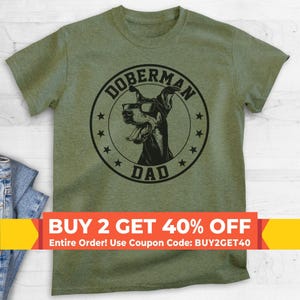Può includere: Una t-shirt a maniche corte color verde oliva con una grafica circolare nera. Il design raffigura un cane Doberman con gli occhiali, circondato da stelle e dalla scritta DOBERMAN DAD. La maglietta ha un girocollo e una vestibilità standard.