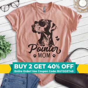 May include: A peach-colored t-shirt with a black and white illustration of a dog wearing sunglasses. The text "Pointer MOM" is printed below the dog. The text "BUY 2 GET 40% OFF" is printed in a yellow and green banner at the bottom of the image.