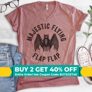 May include: A dusty rose V-neck t-shirt featuring a black graphic of a bat with wings spread wide. The text MAJESTIC FLYING FLAP FLAP is printed in a circular arc around the bat illustration. The shirt has short sleeves and a standard hem.